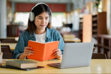Kulaklık takan, kitap okuyan ve kütüphanede dizüstü bilgisayar kullanan odaklanmış bir kadın..
