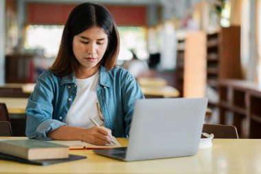 Bir kütüphanede dizüstü bilgisayar ve defterle eğitim ve öğrenmeyi sembolize eden genç bir kadın..