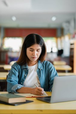 Young woman concentrating on writing notes while using a laptop in a bright study area, showcasing a modern learning environment. clipart