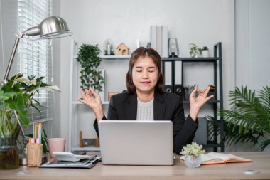 Takım elbiseli bir iş kadını, çevresi bitki ve dizüstü bilgisayarlarla çevrili modern bir ofiste masasında meditasyon yapıyor..