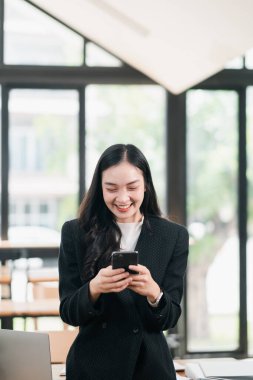 Siyah takım elbiseli profesyonel bir kadın akıllı telefon kullanıyor. Parlak ve modern bir ofis ortamında gülümsüyor..