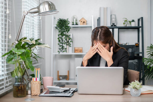 A businesswoman sits at her desk, stressed, with her hands on her face, surrounded by plants and office supplies in a modern office setting.