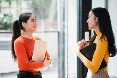 Modern ofis ortamında sohbet eden ve gülümseyen iki genç kadın, ellerinde defterler tutuyorlardı..