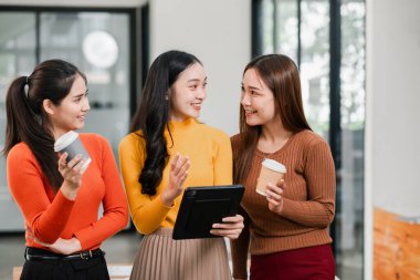 Parlak bir ofiste, elinde kahve fincanları olan, arkadaşça sohbet eden, modern çalışma kültürünü sergileyen üç kadın..