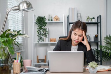 Ev ofisinden genç bir kadın dizüstü bilgisayarla çalışırken stresli görünüyor. Etrafı bitkilerle ve modern dekorlarla çevrili..