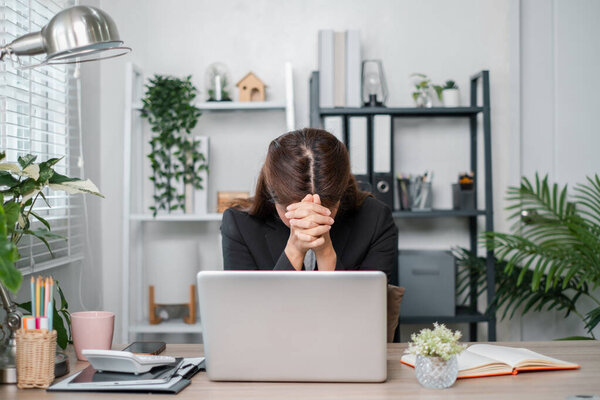 A businesswoman in a modern office, stressed and overwhelmed, sitting at a desk with a laptop, surrounded by plants and office supplies.