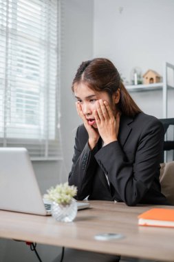 Masasında siyah takım elbiseli bir iş kadını, modern ofis ortamında dizüstü bilgisayarındaki bir şeye tepki veriyor..