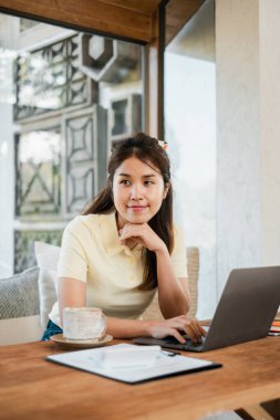 Evde dizüstü bilgisayar kullanan genç bir kadın, elinde kahve fincanı ve not defteriyle pencerenin yanında oturuyor, rahat bir çalışma ortamının keyfini çıkarıyor..