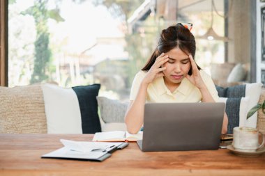 Genç bir kadın evinde dizüstü bilgisayarla çalışırken stresli hissediyor. Evraklar ve rahat bir oturma odasında dizüstü bilgisayarla çevrili..