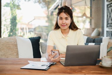 Dizüstü bilgisayarda çalışan, akıllı telefon kullanan, rahat ev dekorlarıyla çevrili, rahat bir kadın..