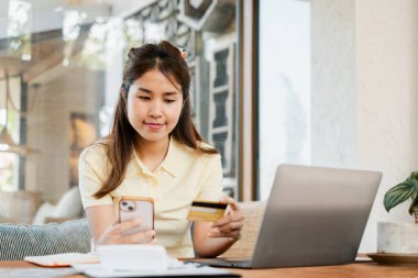 Genç bir kadın rahat bir oturma odasında otururken internet üzerinden alışveriş yapmak için akıllı telefonunu ve kredi kartını kullanıyor..
