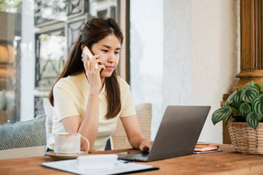 Konforlu bir ofis ortamında dizüstü bilgisayar ve akıllı telefondan birden fazla iş yapan bir kadın, uzaktan çalışma ve iletişimi gösteriyor..
