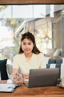 Dizüstü bilgisayar ve akıllı telefon kullanan genç bir kadın rahat ve modern bir oturma odasında doğal ışık altında çalışıyor..