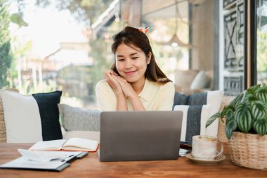 Çiçekler ve kitaplarla çevrili şık bir ev ofisinde dizüstü bilgisayar kullanan gülümseyen bir kadın..