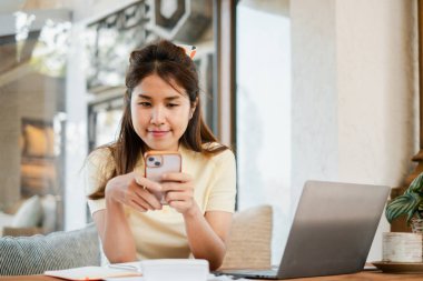 Kafede akıllı telefonunu kullanan genç bir kadın, dizüstü bilgisayarını ve defterini masaya koyup rahat bir çalışma ortamı yaratıyor..