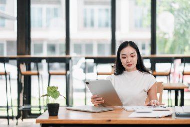 Geniş pencereli, parlak ve modern bir ofiste tablet kullanan genç bir kadın işine odaklanıyor..