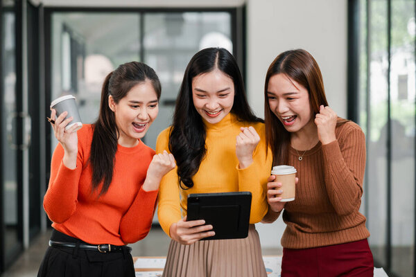 Три девушки в современном офисе радостно празднуют успех, глядя на планшет, держа в руках кофейные чашки.