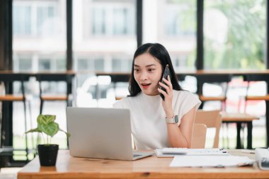 Genç bir kadın, parlak ve modern bir ofis ortamında dizüstü bilgisayarla çalışırken hevesle akıllı telefonuyla konuşuyor..