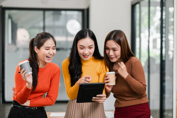 Три девушки в современном офисе, участвуют в командной работе и обсуждениях, пользуются цифровым планшетом и кофе.