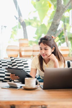 Evde tablet ve laptop kullanan genç bir kadın, doğal ışık ile rahat ve üretken bir uzaktan kumanda düzeneğinin keyfini çıkarıyor..
