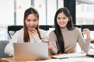 İki genç kadın takım çalışması yapıyor. Parlak ve modern ofis ortamında dizüstü bilgisayar kullanıyorlar..