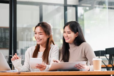 Modern bir ofiste dizüstü bilgisayar ve tablet kullanarak dijital bir projede birlikte çalışan iki genç kadın..