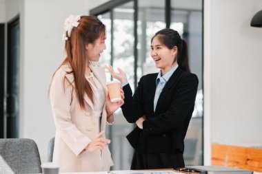 Modern bir ofis ortamında iki iş kadını, kahve ve belgelerle hafif bir anı paylaşıyorlar..