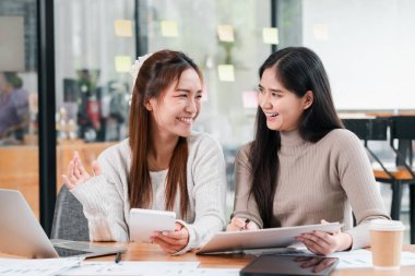 Modern bir ofiste, dijital cihazlarla işbirliği yapan, gülümseyen ve yaratıcı işler yapan iki genç kadın..