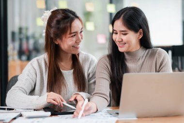 Modern bir ofiste birlikte çalışan iki genç kadın dizüstü bilgisayarlar ve belgeler kullanarak takım çalışması ve işbirliği sergiliyorlar..