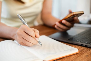Bilgisayarı ahşap bir masanın üstünde, elinde akıllı telefon tutarken deftere yazı yazan birinin yakın plan fotoğrafı..