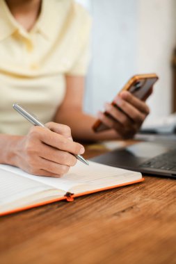 Tahta bir masanın üzerine dizüstü bilgisayarla akıllı telefon tutarken deftere yazı yazan birinin yakın plan fotoğrafı..