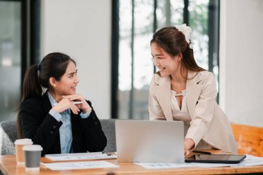 Modern ofis ortamında dizüstü bilgisayarlar ve belgelerle fikirleri tartışan iki kadın..