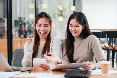 Modern bir ofiste laptoplarla ve belgelerle işbirliği yapan, gülümseyen ve yaratıcı işler yapan iki genç kadın..