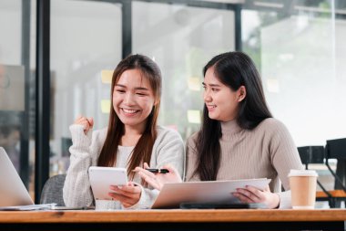 Modern bir ofiste birlikte çalışan iki genç kadın, dizüstü bilgisayarlar ve tabletler kullanarak, takım çalışması ve işbirliği sergiliyorlar..