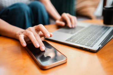 Ev ortamında modern teknoloji kullanımını tasvir eden, ahşap bir masada dizüstü bilgisayar ve akıllı telefon ile etkileşime giren eller..