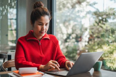 Kırmızı kazaklı bir kadın, yeşil ve doğal ışıkla çevrili, parlak bir kafede dizüstü bilgisayarıyla çalışıyor..