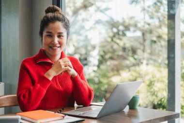 Kırmızı kazaklı, gülümseyen bir kadın bir kafede dizüstü bilgisayarla çalışıyor. Etrafı yeşillik ve doğal ışıkla çevrili..