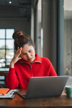 Kırmızı kazaklı odaklanmış bir kadın, doğal ışık ve rahat bir atmosferle çevrili rahat bir kafede dizüstü bilgisayarıyla çalışıyor..