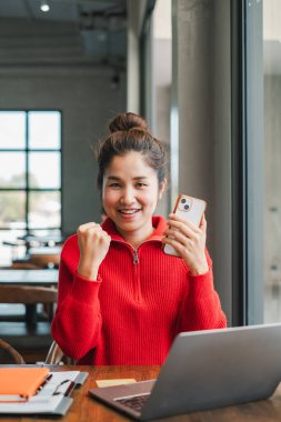 Kırmızı kazaklı neşeli genç bir kadın, elinde akıllı bir telefonla rahat bir kafede dizüstü bilgisayarla otururken başarısını kutluyor..