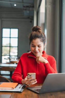 Parlak, modern bir kafede akıllı telefon ve laptop kullanan kırmızı kazaklı, gülümseyen bir kadın..