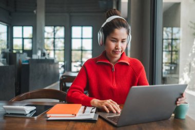 Kırmızı kazaklı bir kadın modern bir kafede dizüstü bilgisayar ve kulaklık kullanıyor. Etrafı defterler ve kahveyle çevrili..