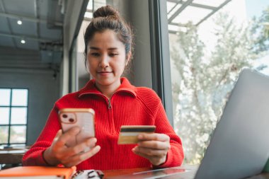 Parlak, modern bir kafe ortamında online alışveriş için akıllı telefon ve kredi kartı kullanan kırmızı kazaklı bir kadın..