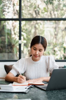 Odaklanmış genç bir kadın, bir ofis masasında dizüstü bilgisayarı ve büyük bir pencereden yayılan doğal ışıkla bir deftere yazar..