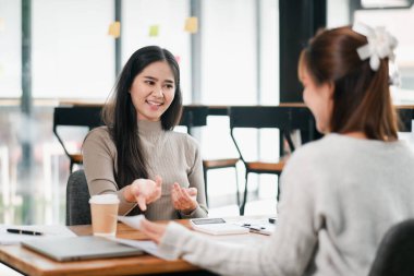 Modern bir kafe ortamında iki kadın iş görüşmesi yapıyor. Masada kahve ve dizüstü bilgisayarlar var..