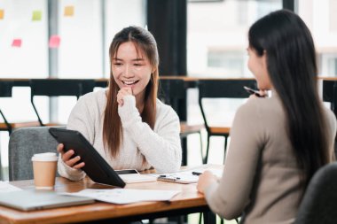 İki genç kadın modern bir ofiste çalışmayı tartışıyor, tablet kullanıyor ve kahveden zevk alıyorlar..