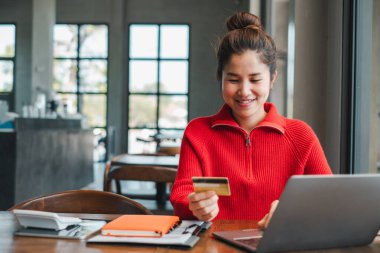 Kırmızı kazaklı genç bir kadın şık bir kafede online alışveriş için dizüstü bilgisayar ve kredi kartı kullanıyor..