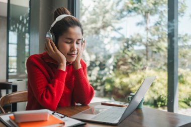 Kırmızı kazaklı genç bir kadın kulaklıkla müzik dinlerken parlak, rahat bir ev ofisinde dizüstü bilgisayarla çalışıyor..