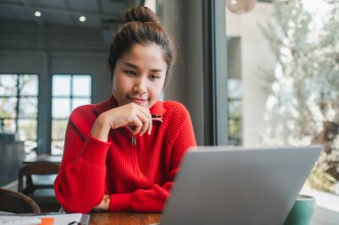Rahat kırmızı kazaklı genç bir kadın geniş pencereli ve yeşillikli, parlak, modern bir kafe ortamında laptopuna odaklandı..