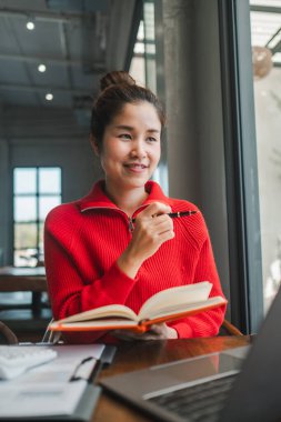 Kırmızı kazaklı, elinde kalem ve defterle gülümseyen bir kadın. Büyük pencerelerden içeri ışık saçan modern bir kafede oturuyor..