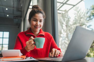 Kırmızı kazaklı, gülümseyen bir kadın elinde kupayla parlak, modern bir ofis alanında dizüstü bilgisayarla çalışıyor..
